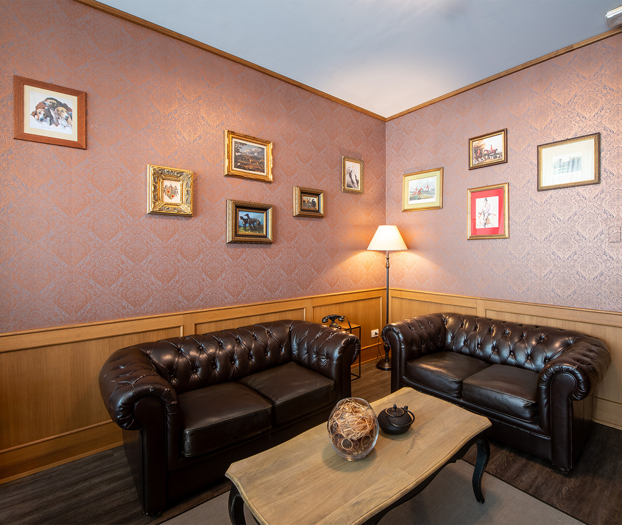 Sala sigari con due divani in pelle scura, tavolino in legno, lampada da terra e parete decorata con quadri incorniciati.