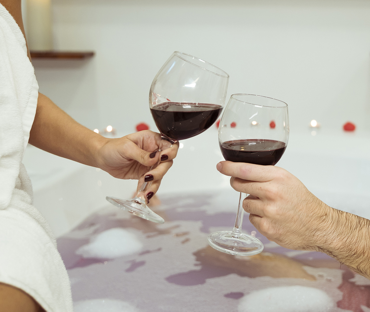 Mani di un uomo e una donna che brindano con calici di vino rosso in una vasca da bagno con schiuma e candele