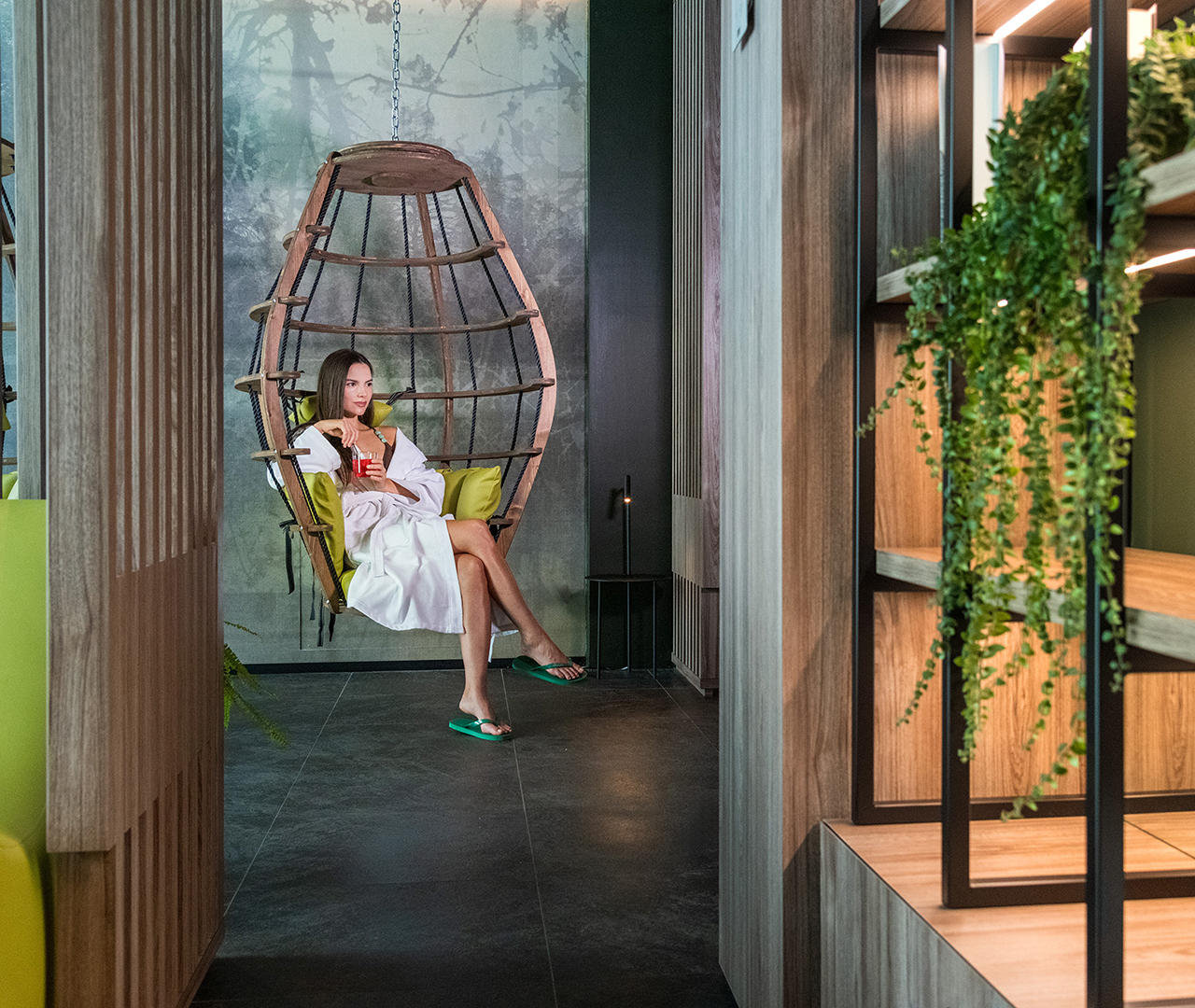 Area relax con poltrona sospesa in legno, cuscini verdi e donna in accappatoio che sorseggia una bevanda.
