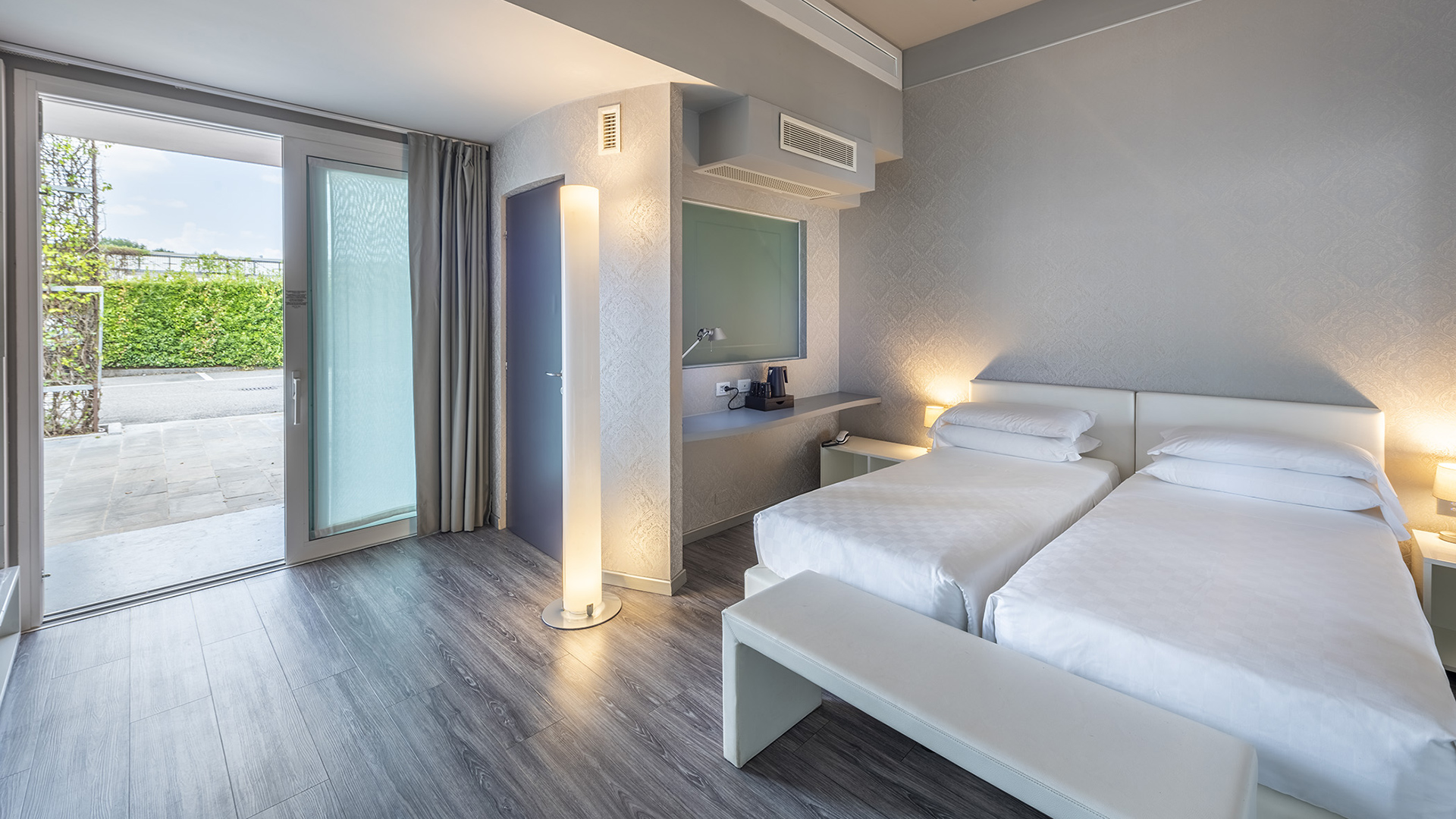 Camera Deluxe con letti singoli, panca ai piedi del letto, scrivania sospesa, lampada da terra e porta finestra con accesso diretto all’esterno