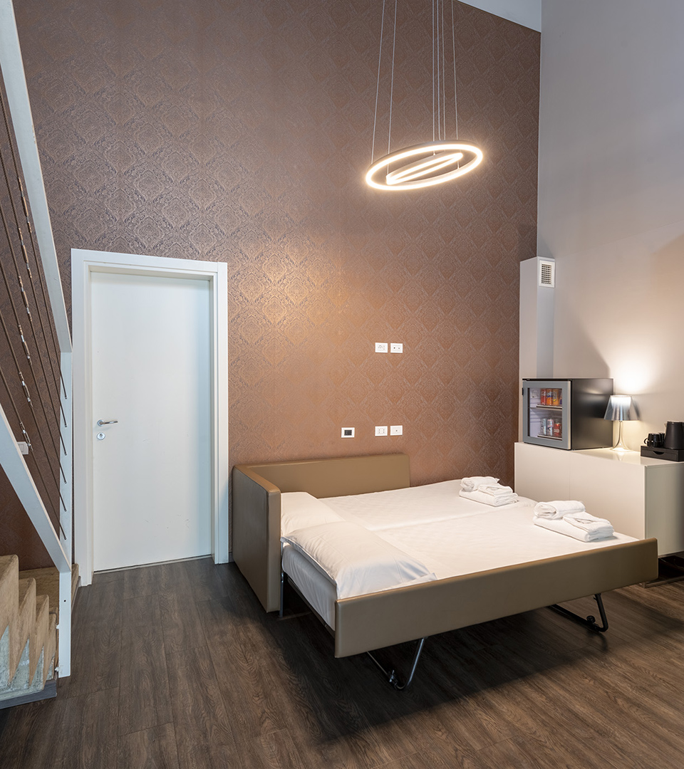 Family Room con letto matrimoniale, parete decorativa color rame, lampada a sospensione circolare, scala interna e angolo con minibar e macchina del caffè