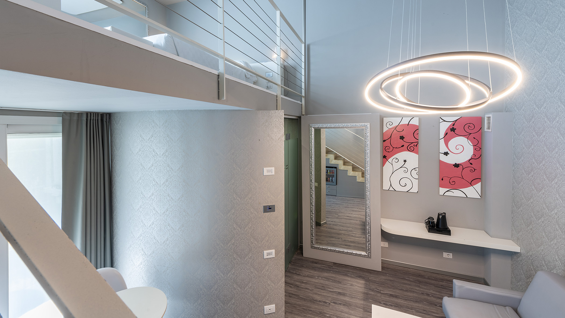 Junior Suite Deluxe su due livelli con soppalco notte, lampada a sospensione circolare di design, grande specchio a parete, scrivania sospesa e area living con poltrona