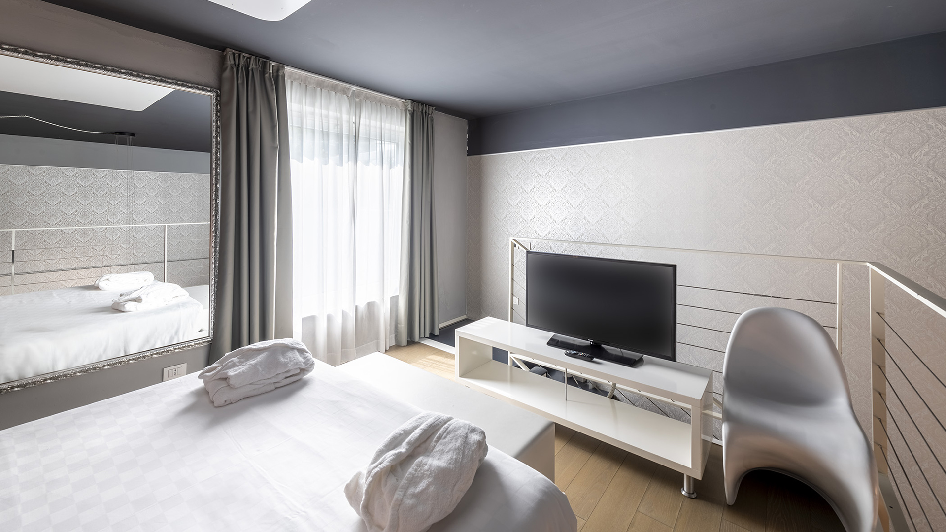 Two-level suite with a double bedroom upstairs, TV on a white cabinet, large wall mirror, window with light curtains, and a designer chair next to the balustrade.