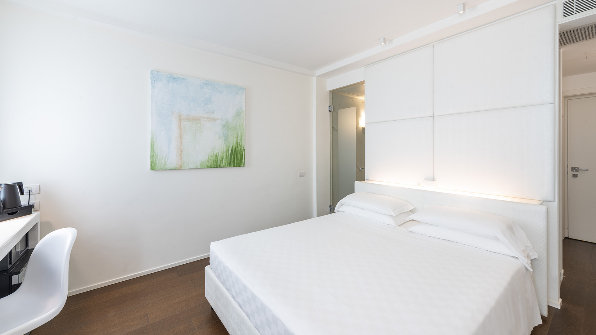 Camera Superior con letto matrimoniale, testiera imbottita bianca con illuminazione integrata, scrivania laterale e pareti minimal con quadro astratto